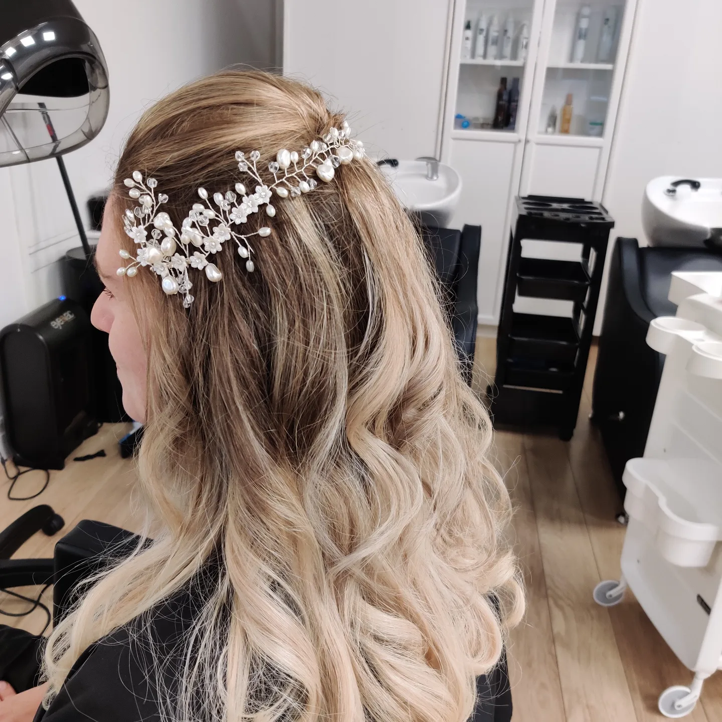 Blonde balayage in krullen met een parelhaarsieraad, bij de wasbakken